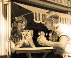Teen boy and girl at a refreshment stand, eating hot dogs and smiling at each other.